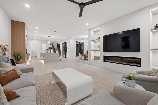 living room featuring built in shelves, recessed lighting, a ceiling fan, stairs, and a glass covered fireplace