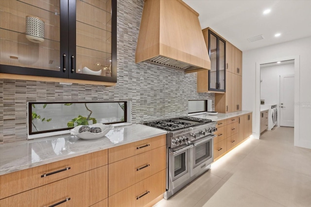 kitchen with tasteful backsplash, custom range hood, glass insert cabinets, light stone countertops, and double oven range