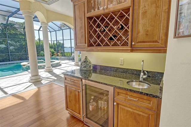 bar with dark stone countertops, sink, beverage cooler, and light hardwood / wood-style flooring