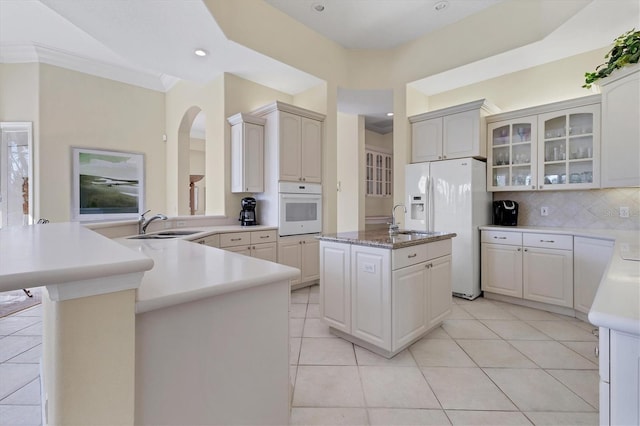 kitchen with a kitchen island with sink, a peninsula, white appliances, light countertops, and glass insert cabinets