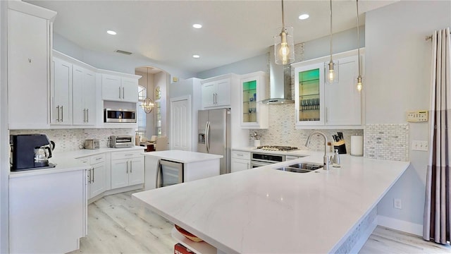 kitchen featuring kitchen peninsula, white cabinets, pendant lighting, stainless steel appliances, and wall chimney range hood