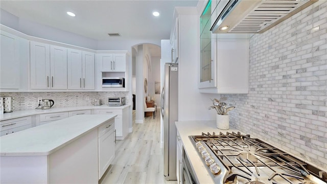 kitchen with wall chimney range hood, appliances with stainless steel finishes, white cabinetry, decorative backsplash, and light wood-type flooring