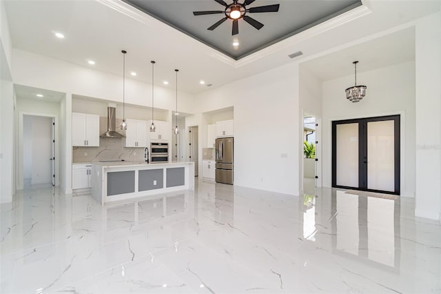 kitchen featuring white cabinets, stainless steel appliances, ceiling fan with notable chandelier, an island with sink, and wall chimney exhaust hood