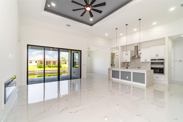 kitchen featuring a tray ceiling, marble finish floor, double oven, white cabinetry, and wall chimney range hood