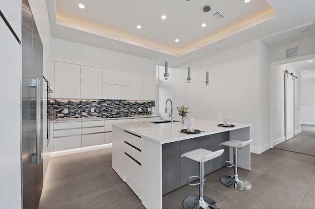 kitchen with an island with sink, white cabinets, a barn door, a tray ceiling, and decorative light fixtures