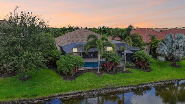 back house at dusk with a water view, a lawn, and a lanai