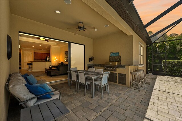 patio terrace at dusk featuring exterior kitchen, ceiling fan, a lanai, a bar, and a grill