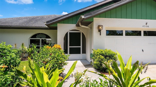 view of exterior entry with a shingled roof, board and batten siding, an attached garage, and stucco siding