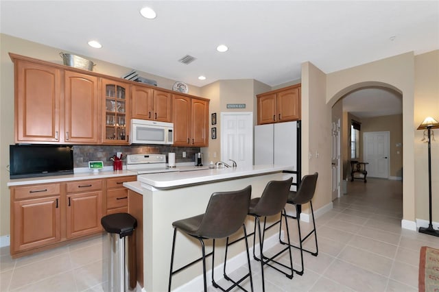 kitchen with a breakfast bar, backsplash, light tile patterned floors, a center island with sink, and white appliances