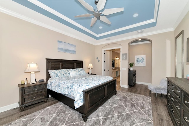 bedroom with crown molding, connected bathroom, ceiling fan, a tray ceiling, and dark hardwood / wood-style floors