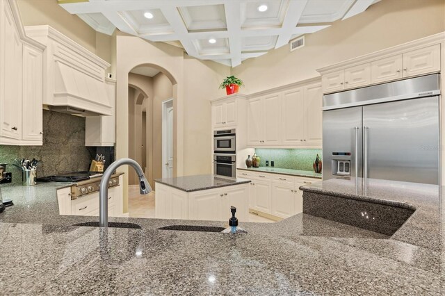 kitchen featuring stainless steel appliances, backsplash, coffered ceiling, dark stone countertops, and a towering ceiling