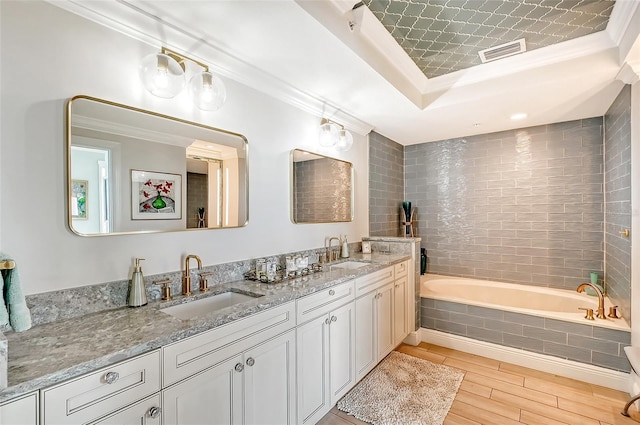 bathroom with wood-type flooring, vanity, a relaxing tiled tub, a tray ceiling, and crown molding