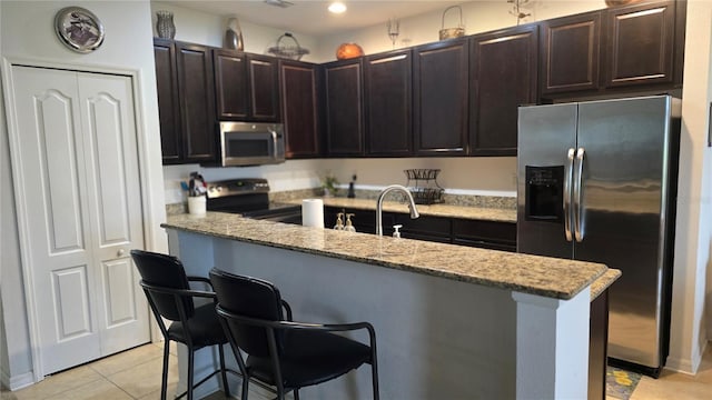 kitchen with light stone countertops, appliances with stainless steel finishes, light tile patterned floors, a kitchen breakfast bar, and a kitchen island with sink