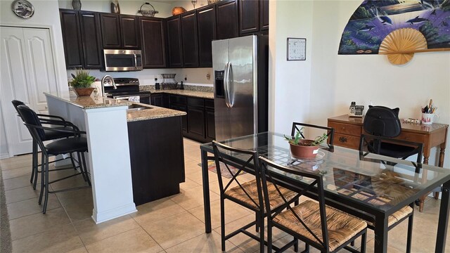 kitchen featuring dark brown cabinetry, a center island with sink, light stone counters, stainless steel appliances, and light tile patterned flooring