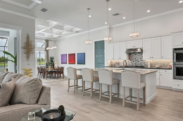 kitchen with a kitchen island with sink, light wood-type flooring, coffered ceiling, and crown molding