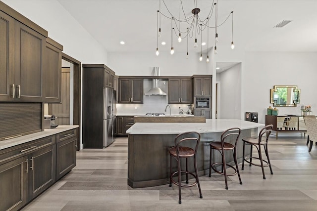 kitchen with a kitchen island with sink, tasteful backsplash, dark brown cabinetry, wall chimney exhaust hood, and light hardwood / wood-style floors