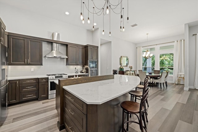 kitchen featuring an inviting chandelier, light stone countertops, a kitchen island with sink, stainless steel appliances, and wall chimney range hood