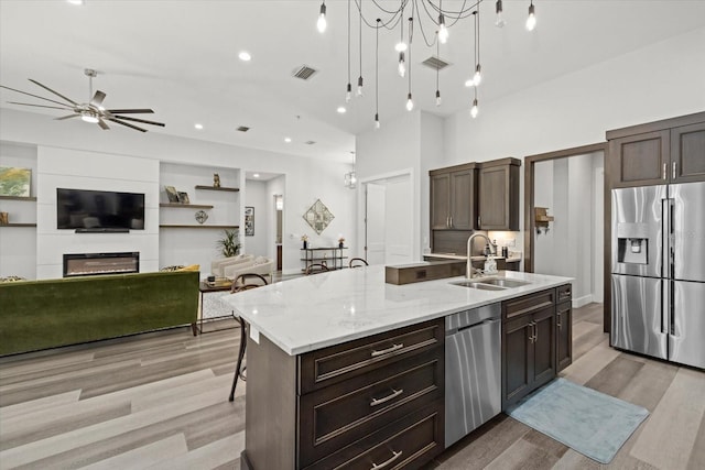 kitchen with ceiling fan with notable chandelier, a fireplace, a kitchen island with sink, stainless steel appliances, and sink