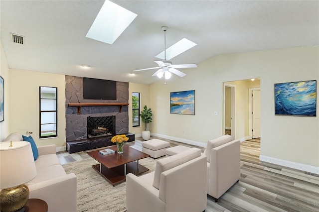 living room featuring light hardwood / wood-style flooring, ceiling fan, a stone fireplace, and vaulted ceiling with skylight