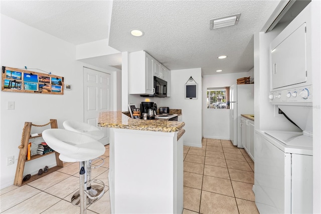 kitchen with white appliances, light tile patterned floors, a textured ceiling, stacked washer and dryer, and white cabinetry