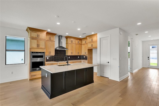 kitchen with wall chimney range hood, sink, light hardwood / wood-style flooring, an island with sink, and double oven