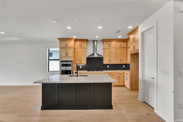 kitchen with appliances with stainless steel finishes, light wood-type flooring, wall chimney exhaust hood, a kitchen island with sink, and light brown cabinets