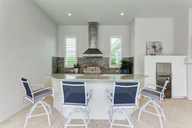 kitchen with decorative backsplash, white cabinetry, stone countertops, wall chimney range hood, and kitchen peninsula