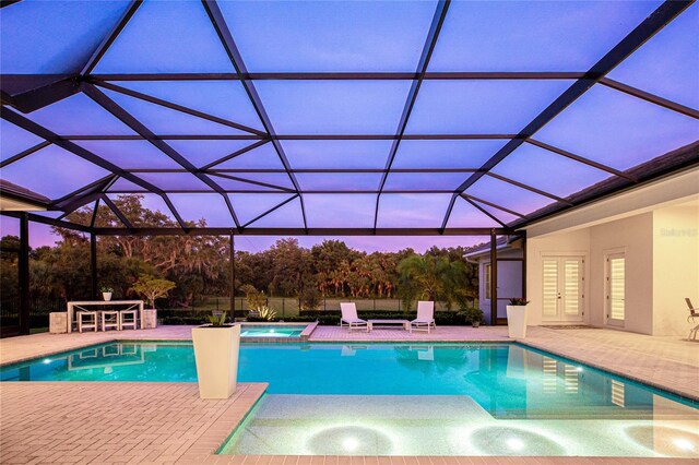 pool at dusk with an in ground hot tub, glass enclosure, and a patio