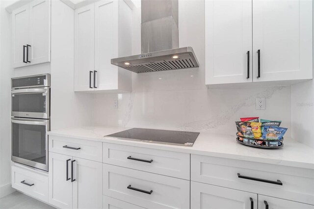 kitchen with white cabinets, stainless steel double oven, black electric stovetop, tasteful backsplash, and wall chimney range hood