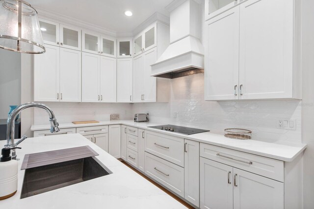 kitchen with black electric cooktop, premium range hood, hanging light fixtures, decorative backsplash, and sink