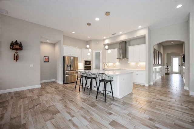 kitchen featuring wall chimney exhaust hood, stainless steel appliances, an island with sink, white cabinets, and pendant lighting