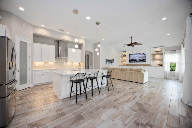 kitchen with a kitchen island with sink, decorative light fixtures, stainless steel refrigerator with ice dispenser, white cabinets, and wall chimney exhaust hood