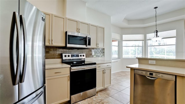 kitchen featuring decorative light fixtures, appliances with stainless steel finishes, a tray ceiling, cream cabinetry, and backsplash