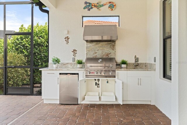 kitchen featuring stainless steel fridge, white cabinetry, light stone counters, sink, and wall chimney range hood