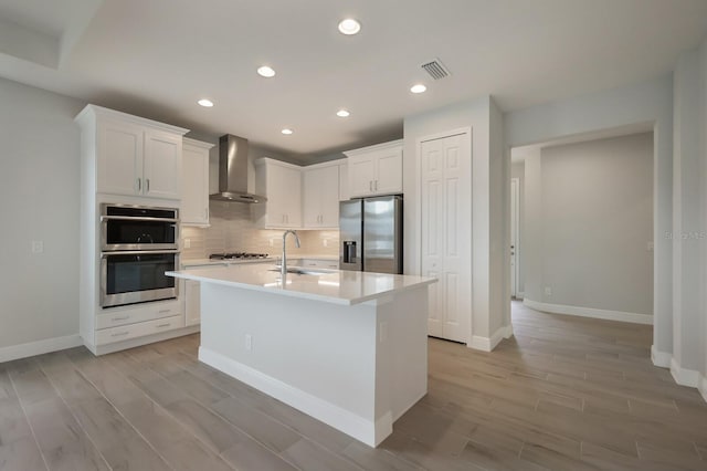 kitchen with sink, a center island with sink, appliances with stainless steel finishes, wall chimney range hood, and white cabinets