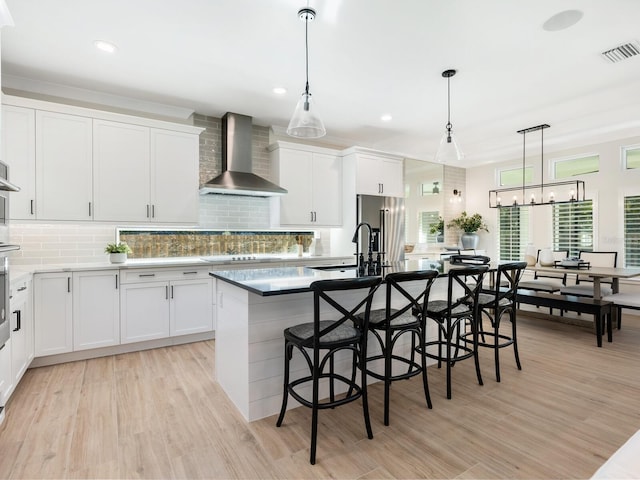 kitchen featuring wall chimney exhaust hood, a breakfast bar, decorative light fixtures, a center island with sink, and white cabinets