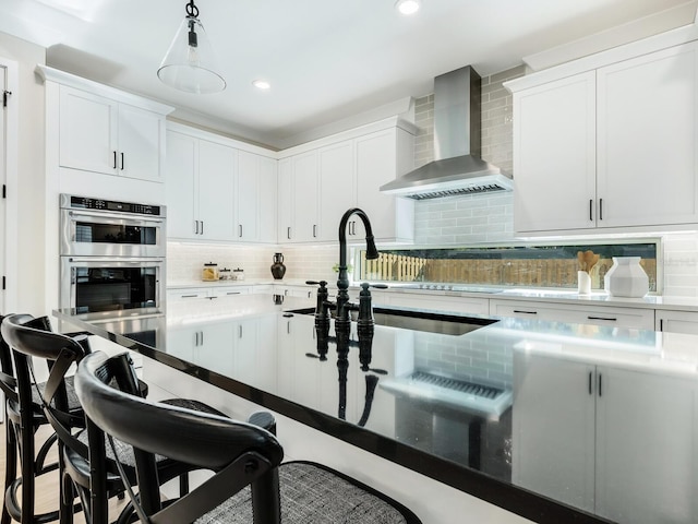 kitchen featuring tasteful backsplash, wall chimney exhaust hood, stainless steel double oven, white cabinets, and a breakfast bar area