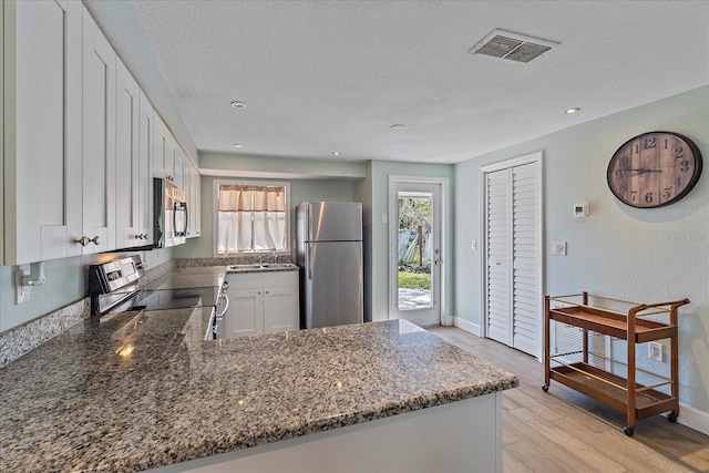 kitchen with a textured ceiling, appliances with stainless steel finishes, light wood-type flooring, dark stone counters, and white cabinetry