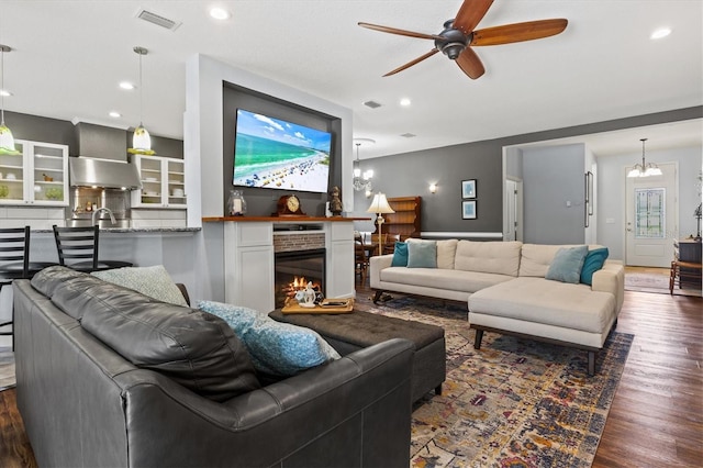 living area featuring recessed lighting, ceiling fan with notable chandelier, visible vents, a lit fireplace, and dark wood-style floors