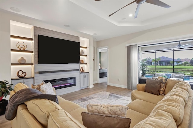 living room with a tray ceiling, built in shelves, ceiling fan, and light hardwood / wood-style flooring