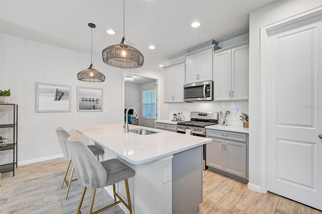 kitchen with appliances with stainless steel finishes, a kitchen island with sink, sink, pendant lighting, and light hardwood / wood-style floors