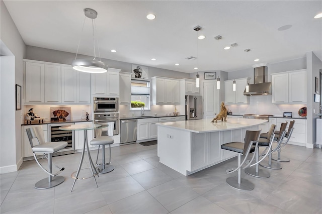 kitchen featuring wall chimney range hood, wine cooler, appliances with stainless steel finishes, a center island, and decorative light fixtures