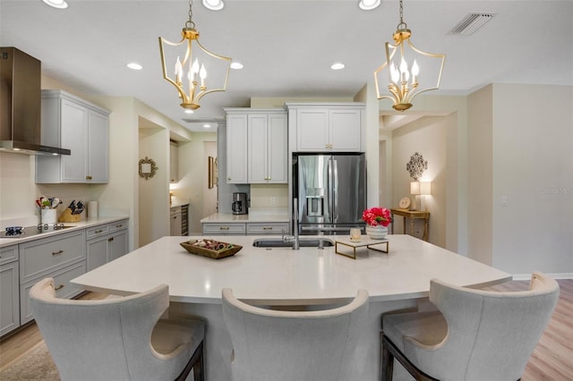 kitchen with an inviting chandelier, black electric cooktop, stainless steel fridge, an island with sink, and wall chimney range hood