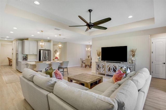 living room with a raised ceiling, ceiling fan with notable chandelier, and light hardwood / wood-style flooring