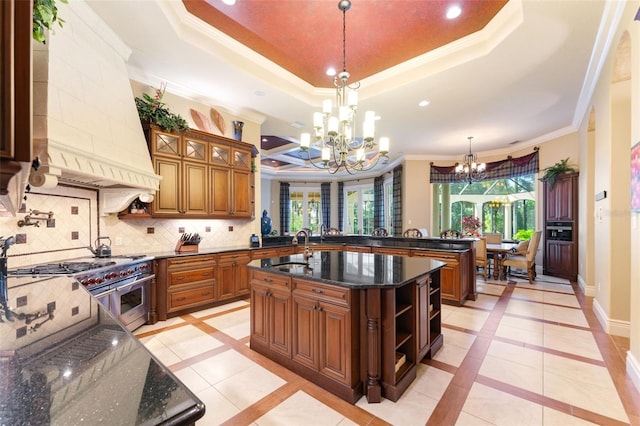 kitchen featuring baseboards, an island with sink, a tray ceiling, premium range hood, and a chandelier