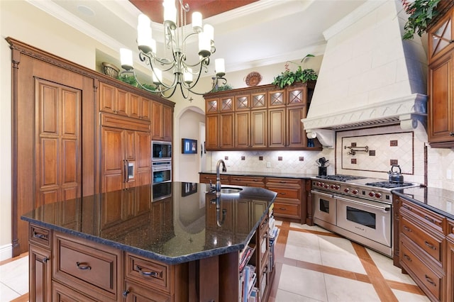 kitchen with backsplash, premium range hood, a center island with sink, light tile patterned floors, and built in appliances