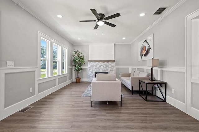 living area featuring dark wood-type flooring, ceiling fan, crown molding, and a stone fireplace