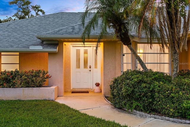 exterior entry at dusk with roof with shingles and stucco siding