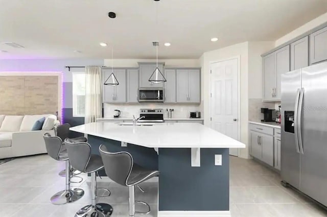 kitchen featuring light tile patterned flooring, appliances with stainless steel finishes, an island with sink, hanging light fixtures, and gray cabinets