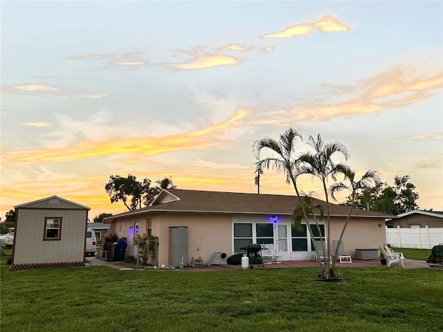 back of house at dusk with an outbuilding, a storage shed, a lawn, stucco siding, and a patio area